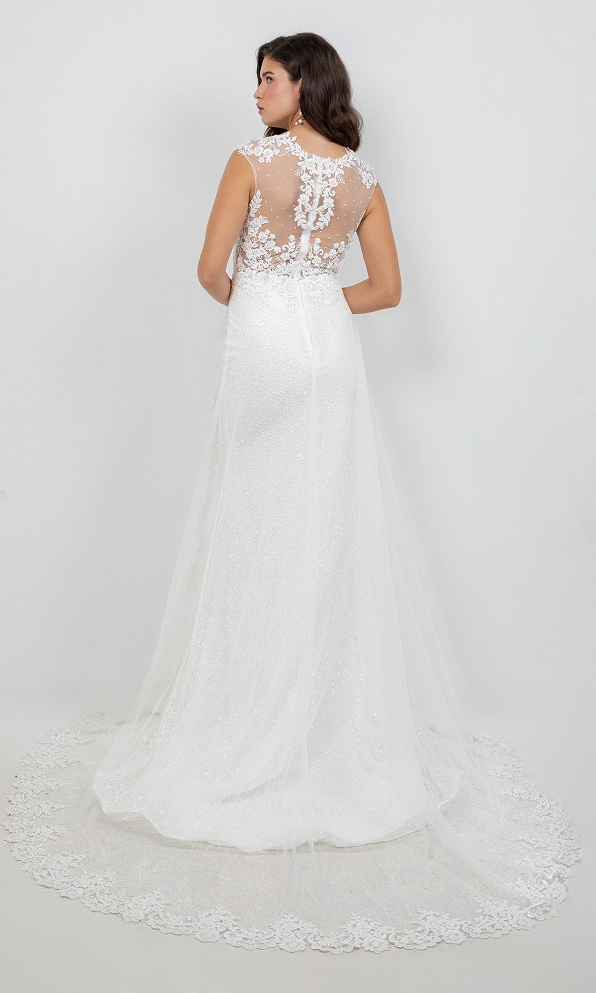 A woman models the Long Formal Dress: Cinderella Couture 8506J, a floor-length white wedding gown featuring lace applique detailing on the back and a sheer tulle train, standing against a plain white background.